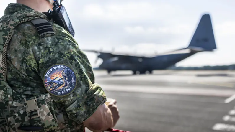 Militair met badge van de oefening op zijn bovenarm staat achter een hekje en kijkt naar een C-130.