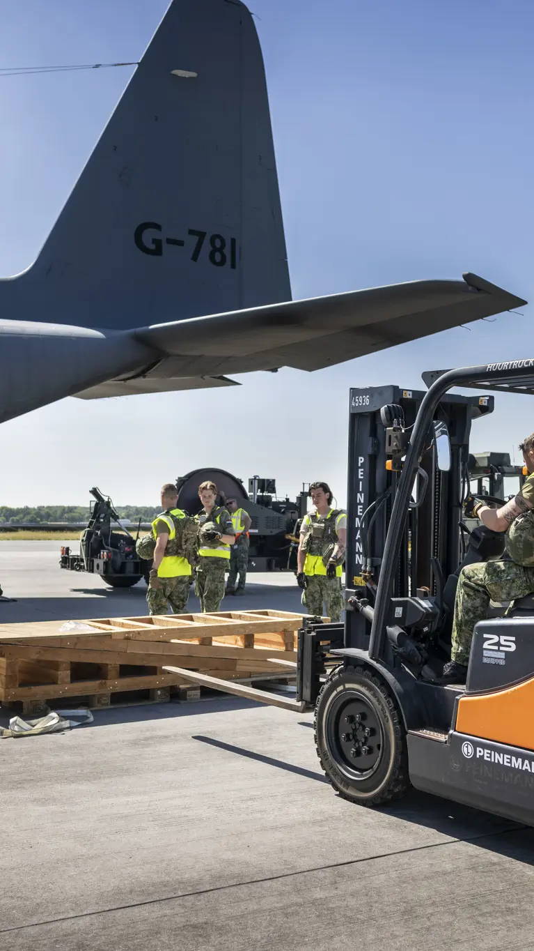 Een vorkheftruck steekt de lepels in een houten stellage. In de achtergrond staat een C-130.