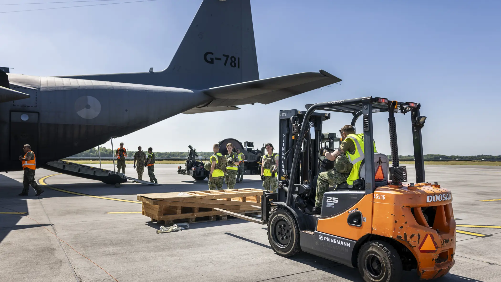 Een vorkheftruck steekt de lepels in een houten stellage. In de achtergrond staat een C-130.