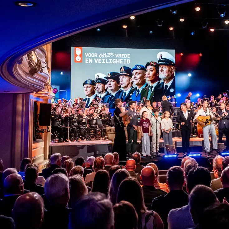 De finale van de ochtend met alle solisten, sprekers, presentatrice en een aantal kinderen op het podium. In de achtergrond is boven het orkest groot een afbeelding van Nederlandse veteranen te zien. 