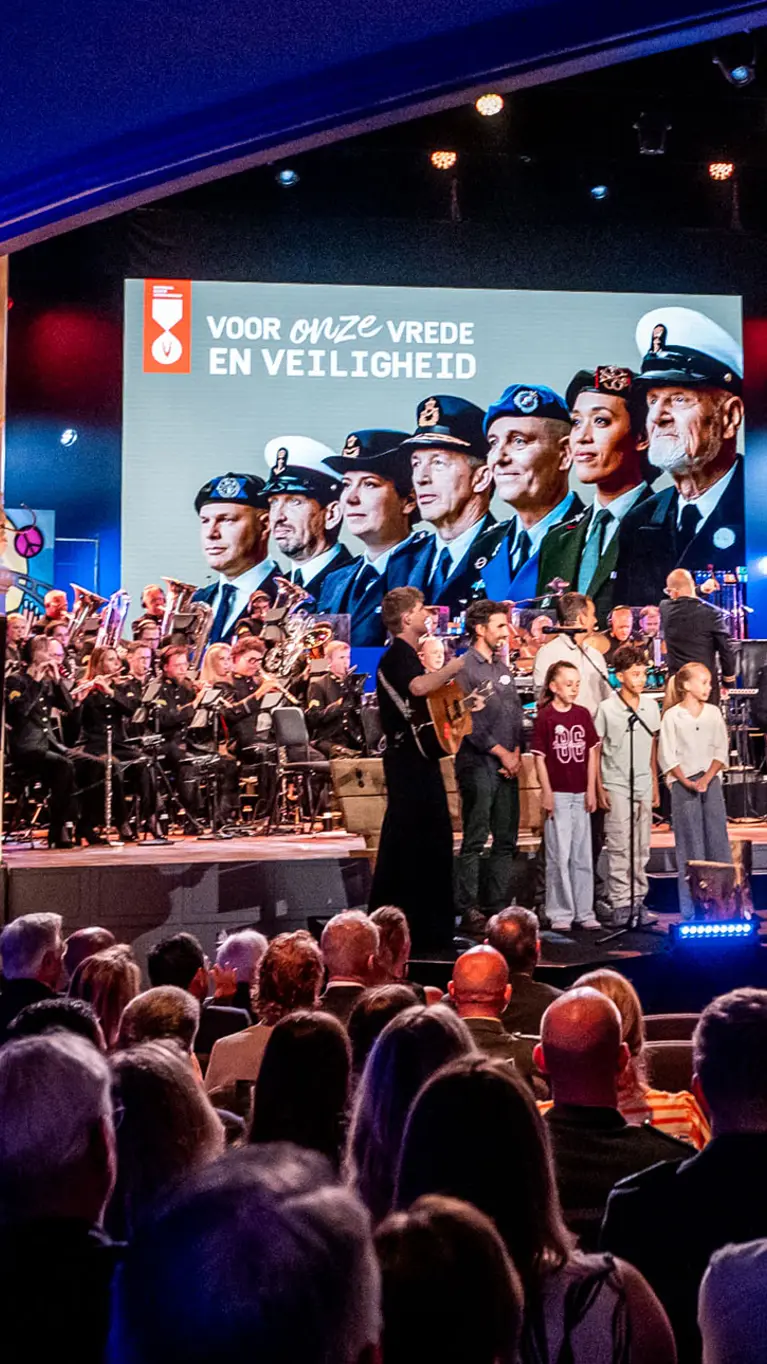 De finale van de ochtend met alle solisten, sprekers, presentatrice en een aantal kinderen op het podium. In de achtergrond is boven het orkest groot een afbeelding van Nederlandse veteranen te zien. 