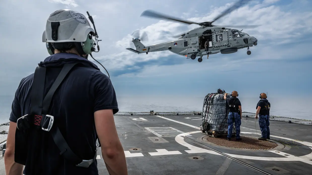 Een helikopter hangt boven het vliegdek terwijl een militair op de voorgrond toekijkt.