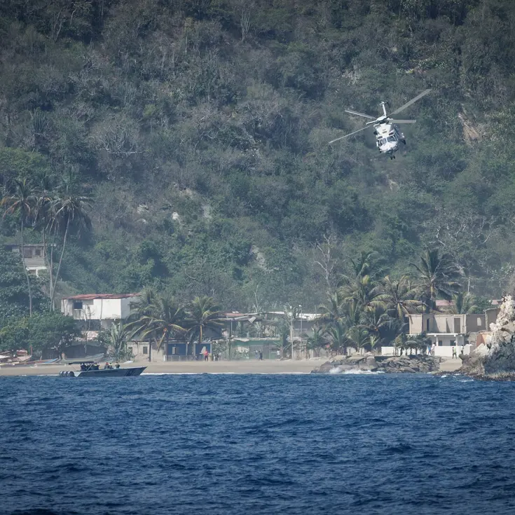 Een helikopter vliegt richting de lens, op de achtergrond groene bergen en wat huisjes.