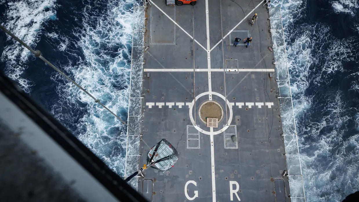 Een pallet met drinkwater wordt opgetild van het vliegdek van Zr.Ms. Groningen, zichtbaar vanuit de helikopter.