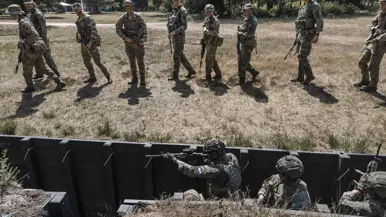 Een man in camouflagekleding wijst naar een groep soldaten met wapens, staand in een open veld met bomen op de achtergrond.