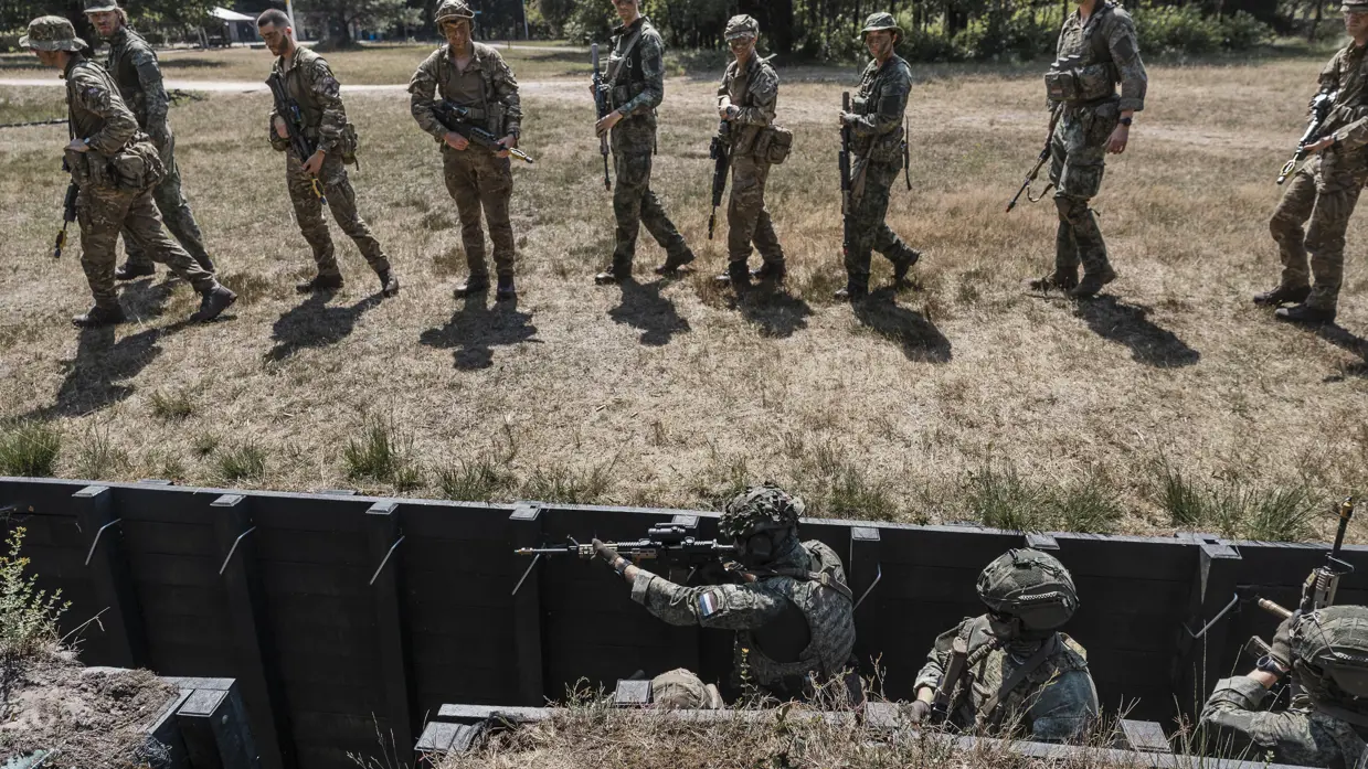 Een man in camouflagekleding wijst naar een groep soldaten met wapens, staand in een open veld met bomen op de achtergrond.