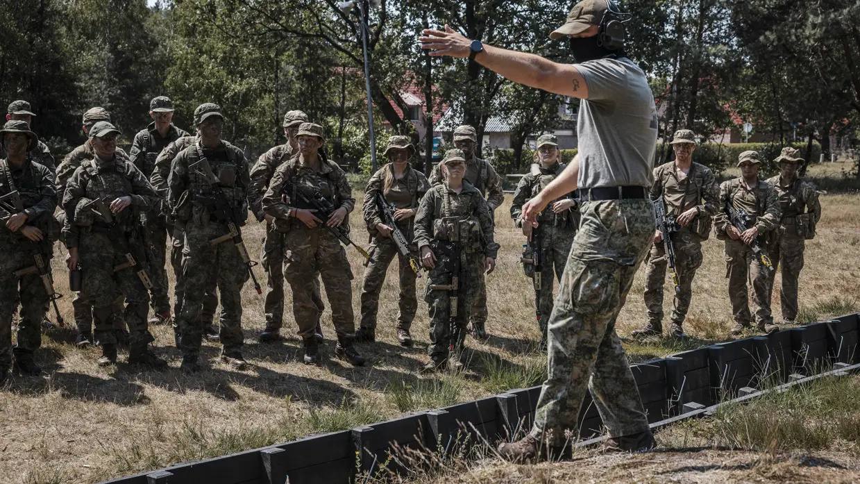 Tien soldaten in camouflagekleding, sommige met wapens, lopen over een open veld; twee liggen achter een zwarte barrière en richten hun wapens.