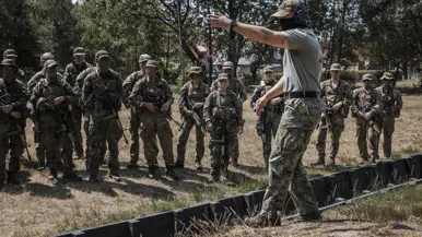 Tien soldaten in camouflagekleding, sommige met wapens, lopen over een open veld; twee liggen achter een zwarte barrière en richten hun wapens.