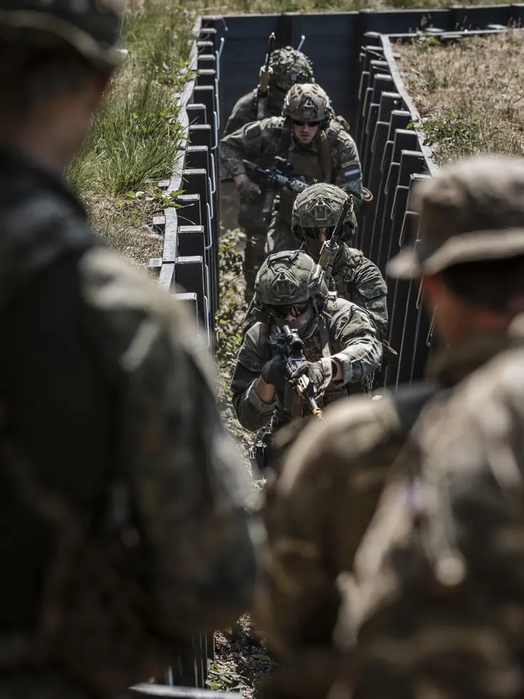 Soldaten in camouflagekleding en helmen, met wapens, klimmen door een smalle loopgraaf in een bosrijk gebied.