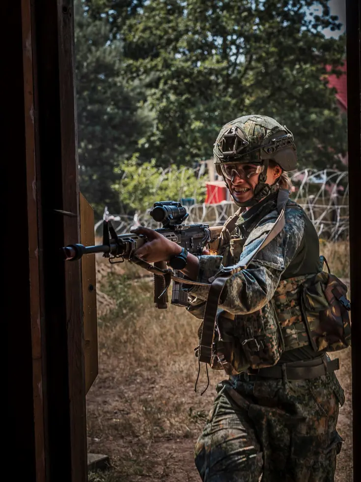 Een soldaat in camouflage met helm en gewapend met een geweer staat alert in een deuropening, omringd door bomen en prikkeldraad.