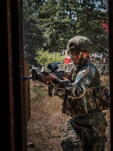 Een soldaat in camouflage met helm en gewapend met een geweer staat alert in een deuropening, omringd door bomen en prikkeldraad.