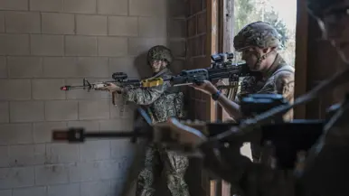 Twee soldaten in camouflage met helmen en geweren richten zich op een doel in een betonnen ruimte met een open deur.