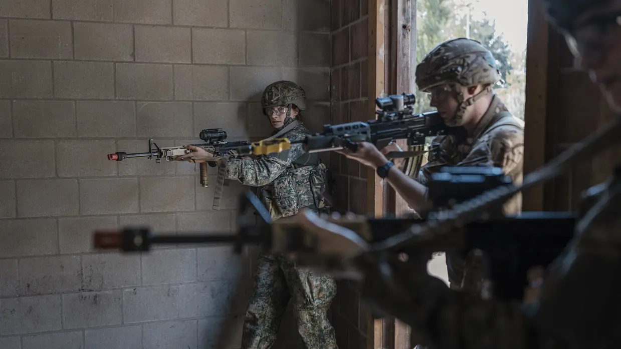 Twee soldaten in camouflage met helmen en geweren richten zich op een doel in een betonnen ruimte met een open deur.