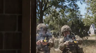 Twee soldaten met helmen en geweren in een deuropening, omringd door bomen en prikkeldraad, in actie.