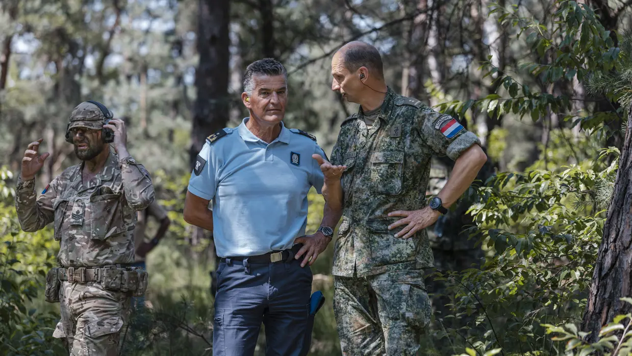 Drie mannen in uniform praten in een bos: twee soldaten in camouflage en één in lichtblauw met insignes.