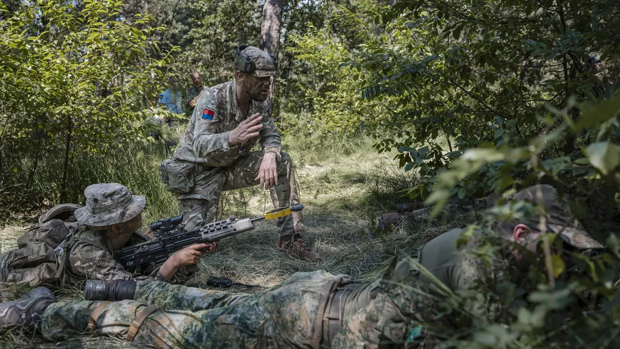 Een militair met een hoed op maakt handgebaren naar twee militairen die op de grond liggen met hun geweer in de aanslag in een beboste omgeving.