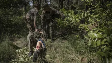 Twee soldaten in camouflage tillen een gewonde collega op een draagbaar door een bos met dichte begroeiing.