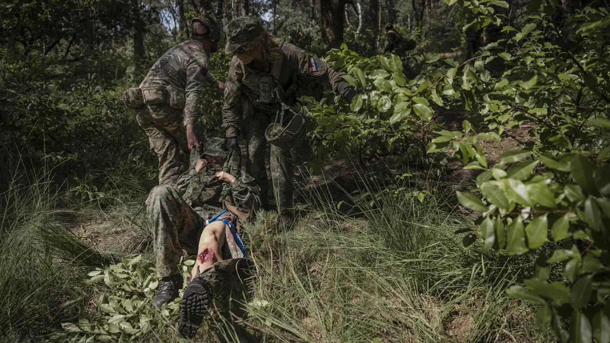 Twee soldaten in camouflage tillen een gewonde collega op een draagbaar door een bos met dichte begroeiing.