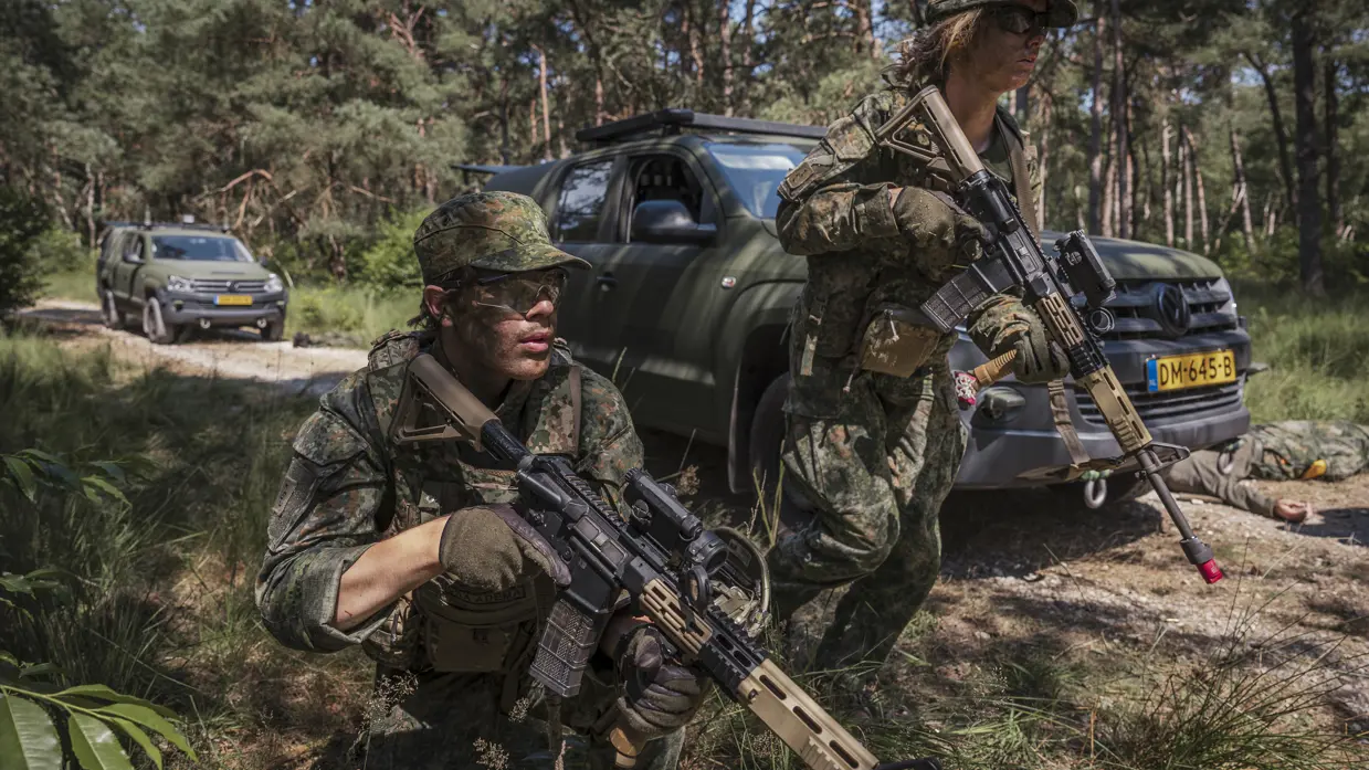 Twee militairen in uniform houden hun wapen vast bij een voertuig in een beboste omgeving.