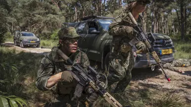 Twee militairen in uniform houden hun wapen vast bij een voertuig in een beboste omgeving.