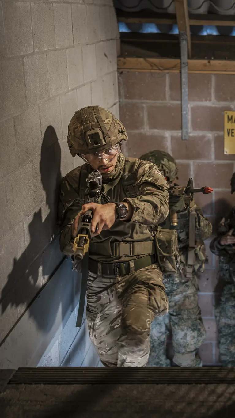 Drie soldaten in camouflage met geweren betreden een donkere betonnen ruimte, alert en gericht op een doel.