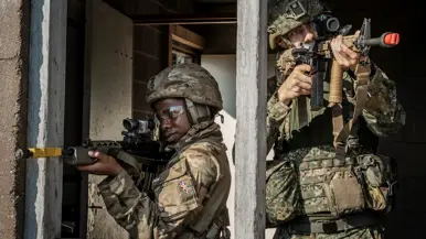 Twee soldaten in camouflage met geweren richten hun wapen door een open raam van een huis Midden:
