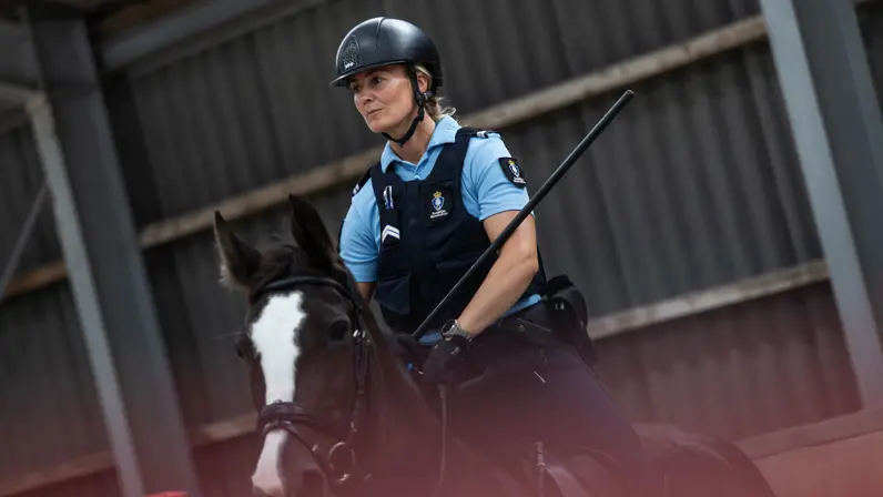 Een vrouw met een blauw marechausseeshirt aan rijdt op een zwart paard. Haar uitgestoken wapenstok raakt een staander met kussens erop.
