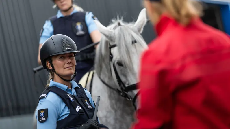 Een dame draagt een blauw marechausseeshirt en zit op een wit paard. Ze kijkt naar de instructeur die vlak voor de cameralens staat. 