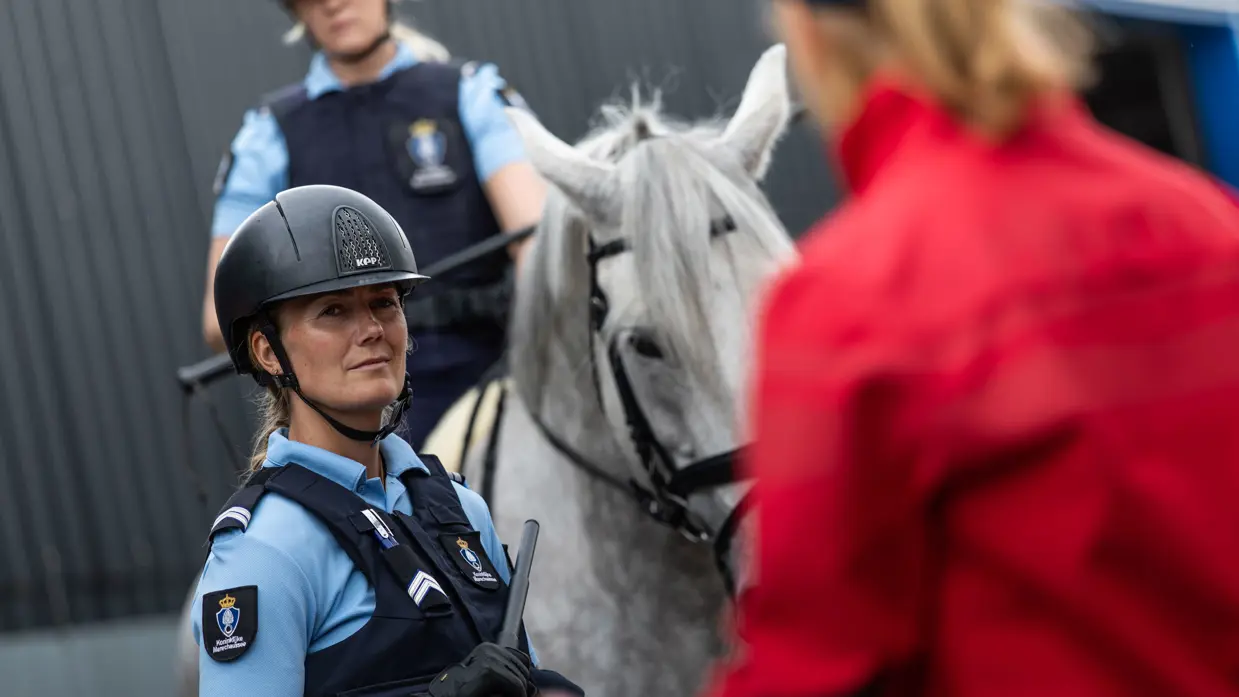 Een dame draagt een blauw marechausseeshirt en zit op een wit paard. Ze kijkt naar de instructeur die vlak voor de cameralens staat. 