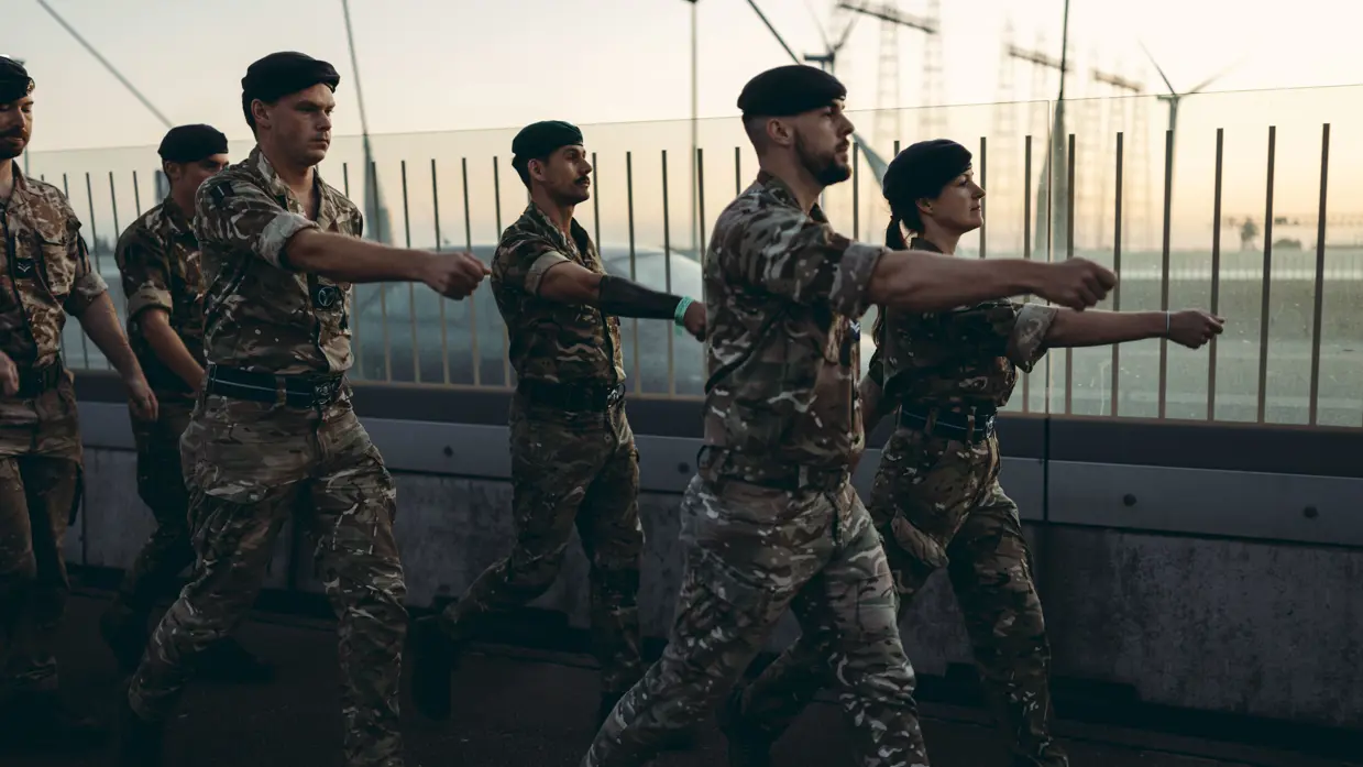 Een groep militairen marcheert over een brug met gestrekte armen.