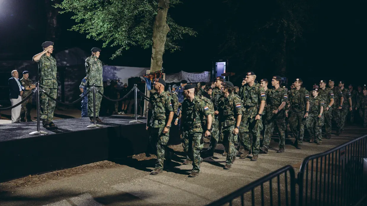 Een groep militairen marcheert in het donker langs een podium, waar 2 groetende militairen op staan.&nbsp;