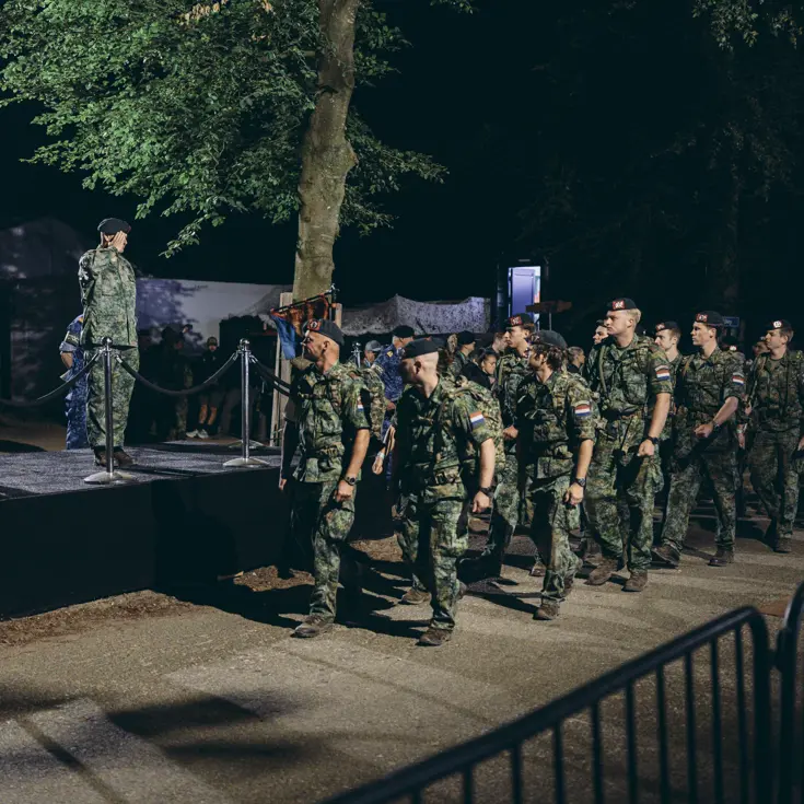 Een groep militairen marcheert in het donker langs een podium, waar 2 groetende militairen op staan.&nbsp;