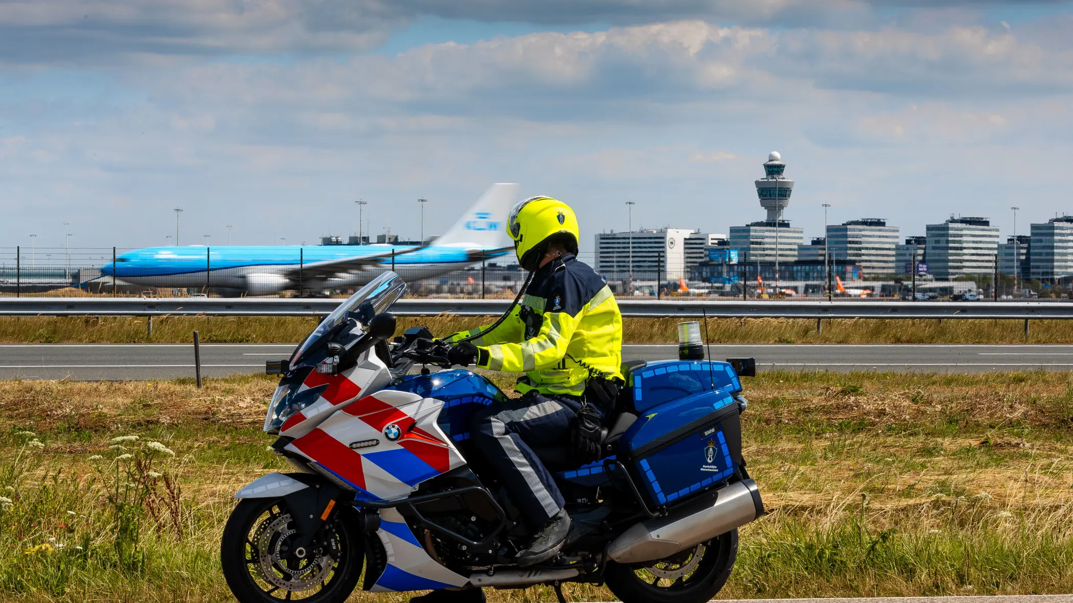 Motorrijder van de Marechaussee, op de rug gezien met Schiphol op de achtergrond