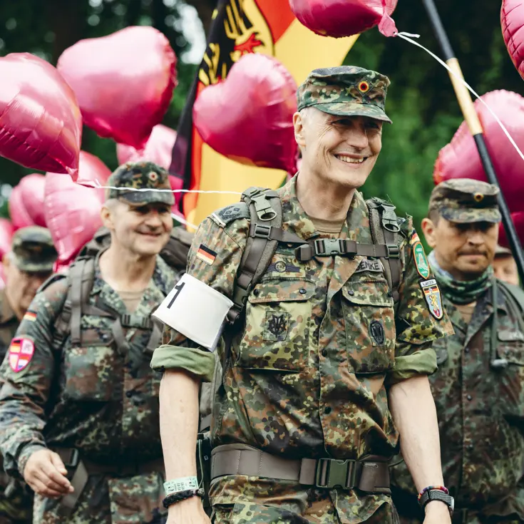 Een groep Duitse militairen marcheert lachend voorbij. Boven hen torenen roze ballonnen.  