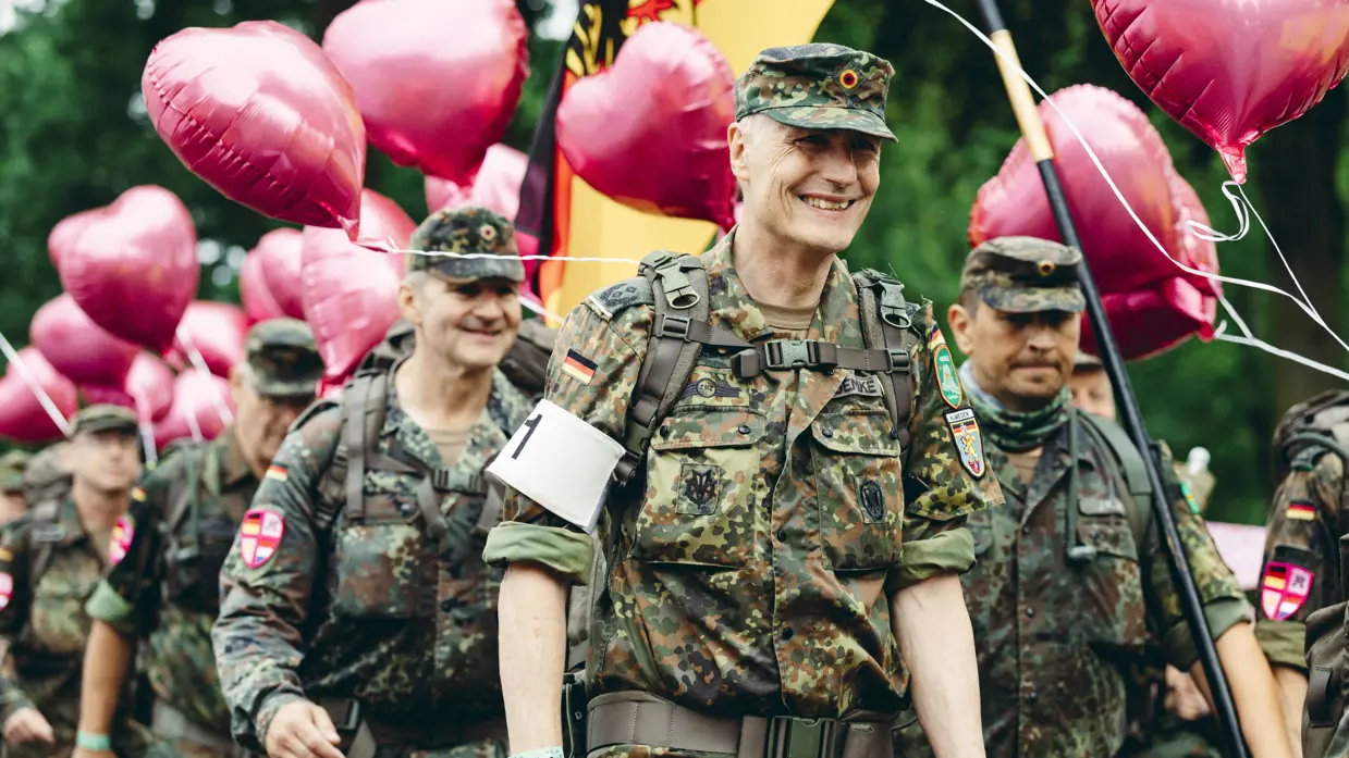 Een groep Duitse militairen marcheert lachend voorbij. Boven hen torenen roze ballonnen.  