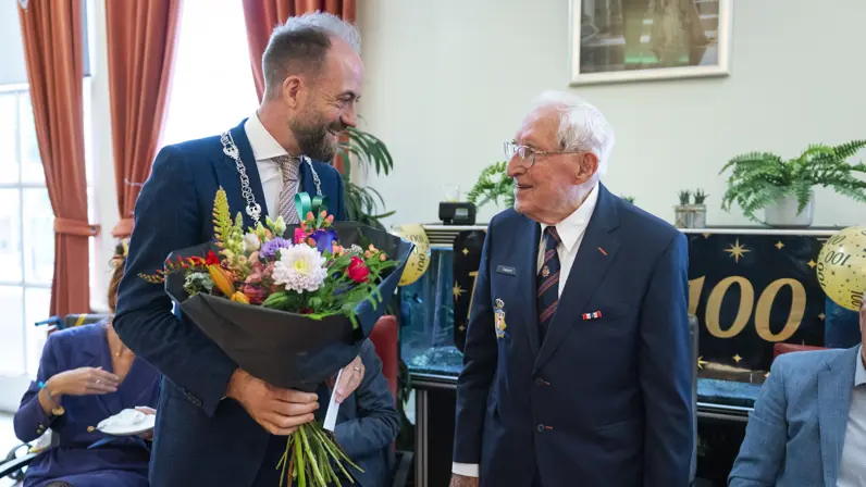Een wethouder overhandigt een bos bloemen aan een honderdjarige veteraan van Bronbeek tijdens zijn verjaardagsviering. 