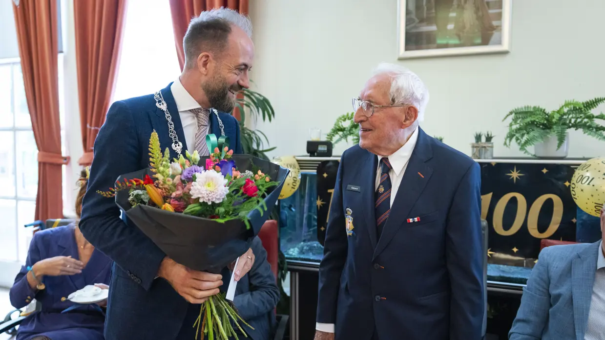 Een wethouder overhandigt een bos bloemen aan een honderdjarige veteraan van Bronbeek tijdens zijn verjaardagsviering. 
