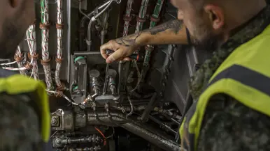 Twee militairen met een geel hesje sleutelen in een technische ruimte van een vliegtuig. 