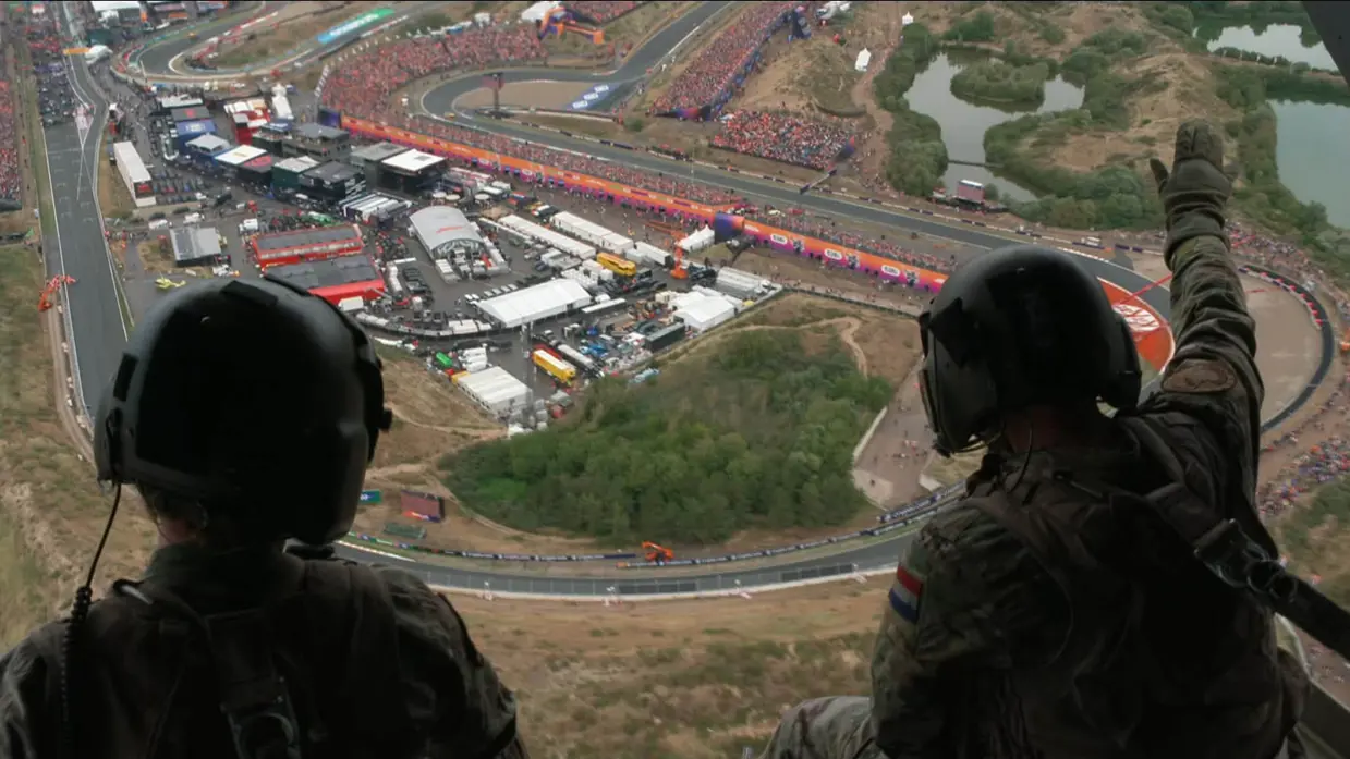 Twee militairen op de rug gezien, zitten op de achterklep van een Chinook (de klep en heli zijn daarbij niet in beeld), vliegend over het Circuit van Zandvoort. Een van de militairen zwaait naar het publiek op de grond.