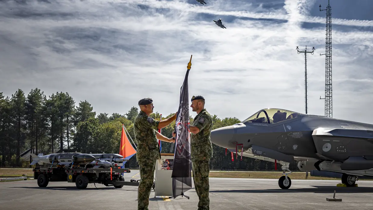 Windhouwer overhandigt aan Kerkhofs een vlag. Op de achtergrond de neus van een F-35 en een aanhanger met daarop F-35 munitie. Er vliegen twee F-35’s over.