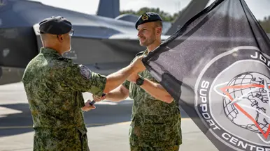 Kerkhofs neemt een vlag van Windhouwer in ontvangst. De vlag met daarop het embleem en de tekst ‘Joint Target Support Centre’ is daarbij half in beeld. Op de achtergrond staat een F-35.