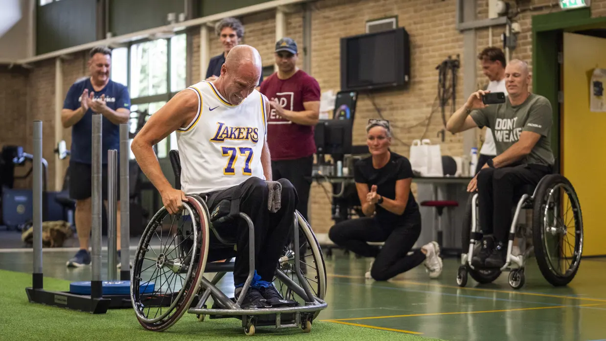 Man in sportoutfit rijdt in een sportrolstoel in een gymzaal onder aanmoediging van vijf mensen, onder wie één in een rolstoel.