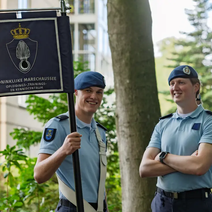 Twee marechaussees lachen naar de camera. De linker heeft een vaandel in de hand.