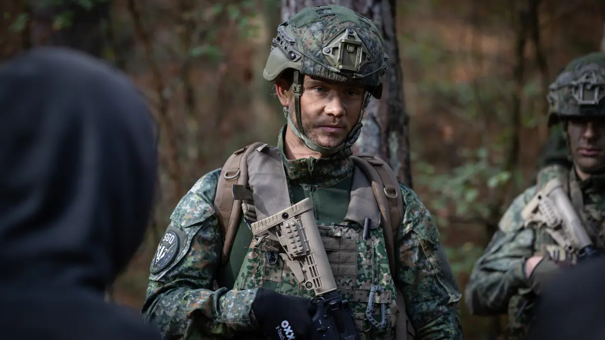 Een militair met stoppelbaard kijkt serieus voor zich uit. In zijn handen een wapen. Een blonde, vrouwelijke militair kijkt opzij en wijst.&nbsp;