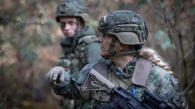 Een militair met stoppelbaard kijkt serieus voor zich uit. In zijn handen een wapen. Een blonde, vrouwelijke militair kijkt opzij en wijst.&nbsp;