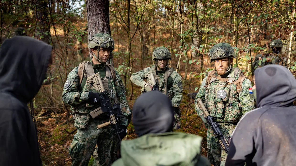 Drie gewapende militairen in gesprek met drie personen in donkere burgerkleding. Een vrouw met een donkere trui en een bedekt gezicht. Militairen in gevecht met de donker geklede partizanen. Twee militairen houden een persoon in burgerkleding vast terwijl diegene met zijn buik op de grond ligt.