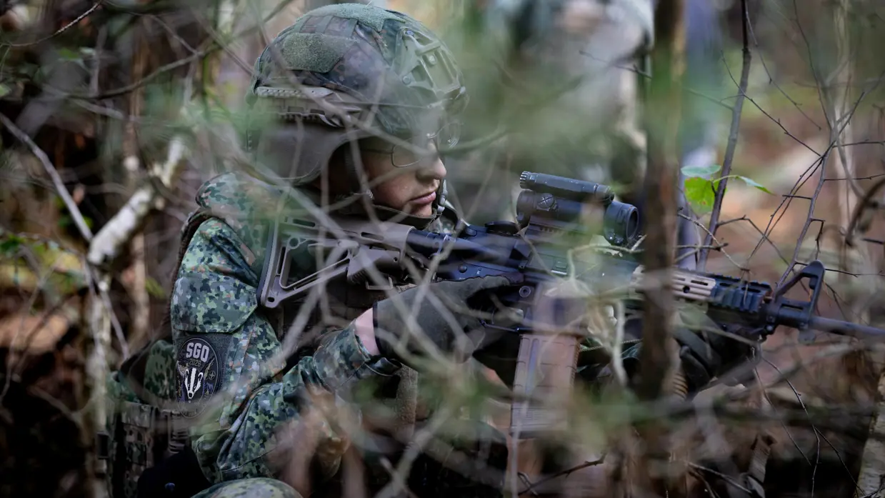 Een geknielde militair met bril op houdt zijn wapen vooruit. 