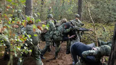 Drie gewapende militairen in gesprek met drie personen in donkere burgerkleding. Een vrouw met een donkere trui en een bedekt gezicht. Militairen in gevecht met de donker geklede partizanen. Twee militairen houden een persoon in burgerkleding vast terwijl diegene met zijn buik op de grond ligt.