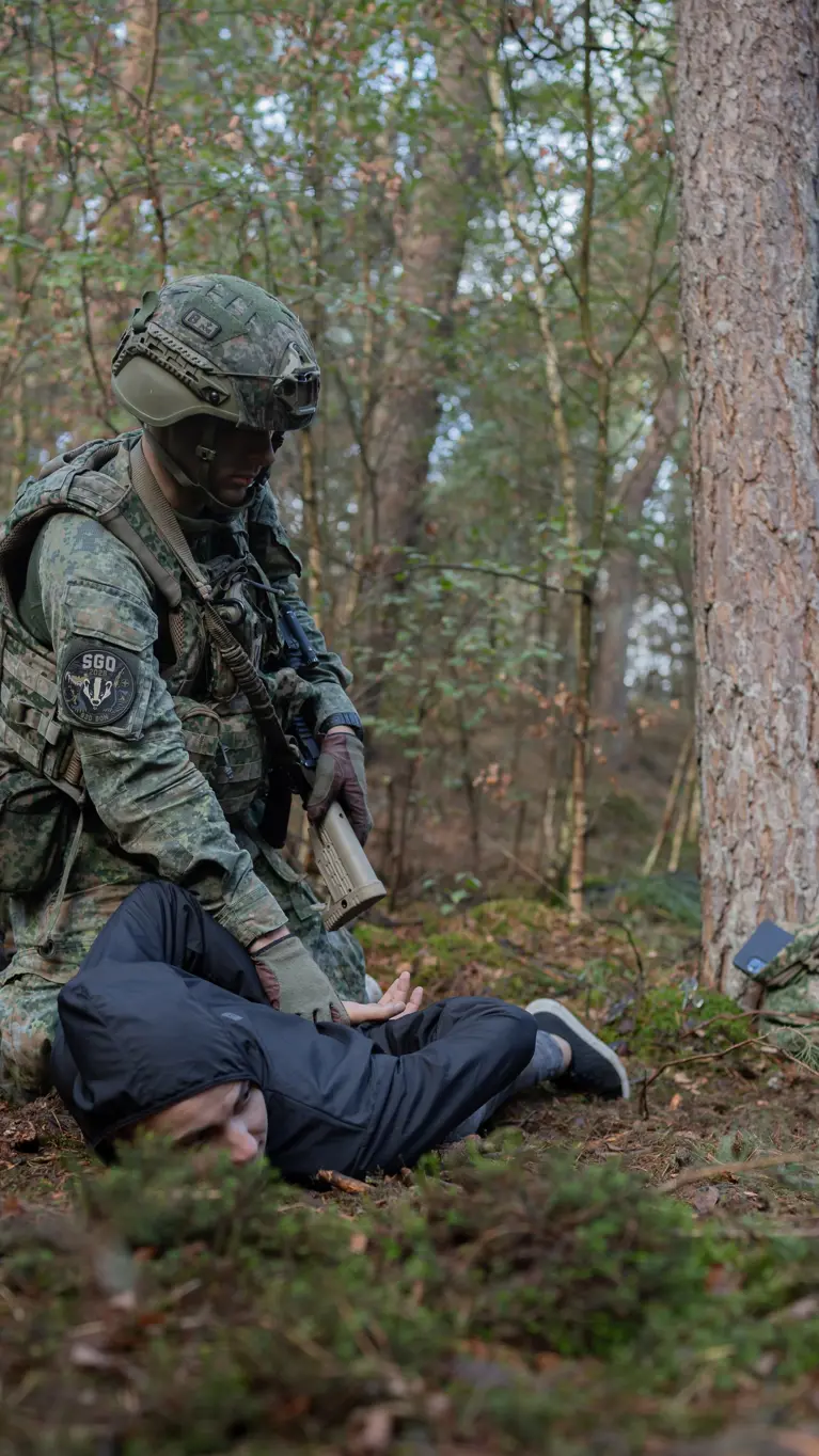  Twee militairen in het bos houden ieder een persoon in burgerkleding tegen de grond gedrukt.