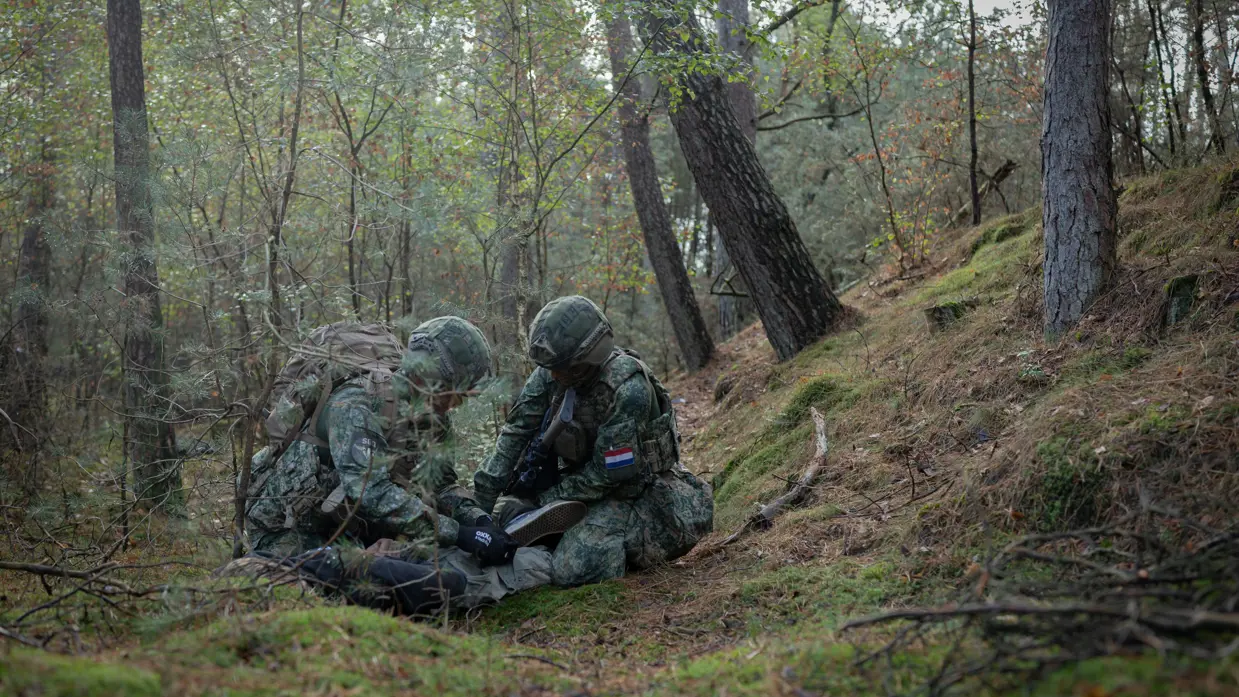 Drie gewapende militairen in gesprek met drie personen in donkere burgerkleding. Een vrouw met een donkere trui en een bedekt gezicht. Militairen in gevecht met de donker geklede partizanen. Twee militairen houden een persoon in burgerkleding vast terwijl diegene met zijn buik op de grond ligt.